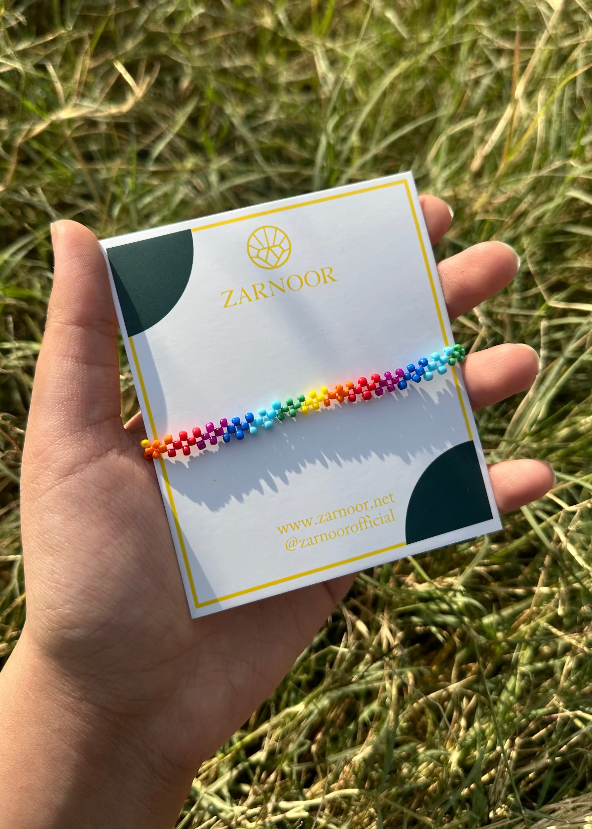 Hand holding a rainbow bracelet on a Zardozi branded card against a grassy background
